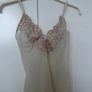 Beige linne med spetsdetaljer - Säljer ett elegant beige linne med vackra spetsdetaljer i rosa och beige runt halsringningen. Linnet har smala axelband och en mjuk, flytande passform. Passar storlek: XS/S