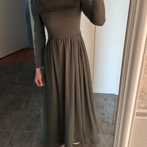 Mörkgrön långklänning med volangdetaljer - Säljer en elegant mörkgrön långklänning med fina volangdetaljer vid bröstet. Klänningen har långa ärmar och en vacker, flytande kjol som ger en klassisk look. Är 166