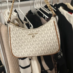 Beige axelväska från Michael Kors - Snygg beige axelväska från Michael Kors med klassiskt MK-mönster. Väskan har en guldfärgad kedja som axelrem och bruna detaljer. Perfekt för både vardag och fest! Väskan är använd 3 gånger. 