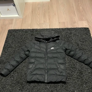 Svart dunjacka från Nike - Säljer en svart dunjacka från Nike i storlek 147-158. Jackan har en huva och dragkedja framtill,  perfekt för kyliga dagar. Den är i bra skick och passar både höst och vinter. En stilren och funktionell jacka för alla tillfällen.