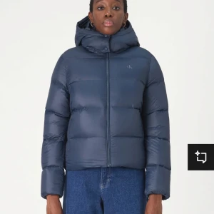 Mörkblå dunjacka med huva - Säljer denna dunjacka från Calvin Klein som är helt oanvänd med prislappen kvar, men som inte passade som jag ville tyvärr. Den är helt slutsåld i denna storleken!