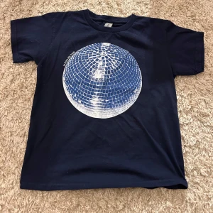 Svart t-shirt med discokultryck från Gildan - Säljer en svart t-shirt från In Print We Trust i bomull med ett coolt discokultryck på framsidan. Perfekt för en avslappnad stil eller en utekväll. T-shirten har korta ärmar och en normal passform. Texten 'In Print We Trust' finns tryckt bredvid discokulan.