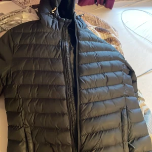 Svart dunjacka med huva - Säljer en svart dunjacka i mycket bra skick. Den har en praktisk huva och dragkedja framtill. Perfekt för kalla vinterdagar. Jackan är stilren och varm, idealisk för både vardag och utomhusaktiviteter.