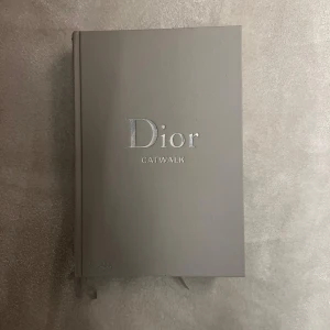 Dior Catwalk - Säljer en stilren och elegant bok med titeln 'Dior Catwalk' från Thames & Hudson. Boken har ett grått omslag med silvertext och innehåller detaljerade beskrivningar av John Gallianos verk. Perfekt för modeintresserade och samlare!