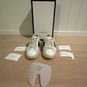 Vita sneakers från Gucci - Säljer ett par vita Gucci sneakers i skinn med den ikoniska röd-gröna randen och en broderad guldfärgad bi-detalj på sidan. Skorna har snörning och är i bra skick, med originalförpackning och tillbehör. Perfekta för både vardag och fest!