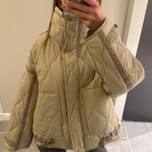Beige quiltad jacka - Säljer en superfin beige quiltad jacka med hög krage och dragkedja. Den har en loose passform och är perfekt för höst och vår. Jackan har långa ärmar och praktiska fickor framtill. Perfekt för att hålla sig varm och stilren! 💫