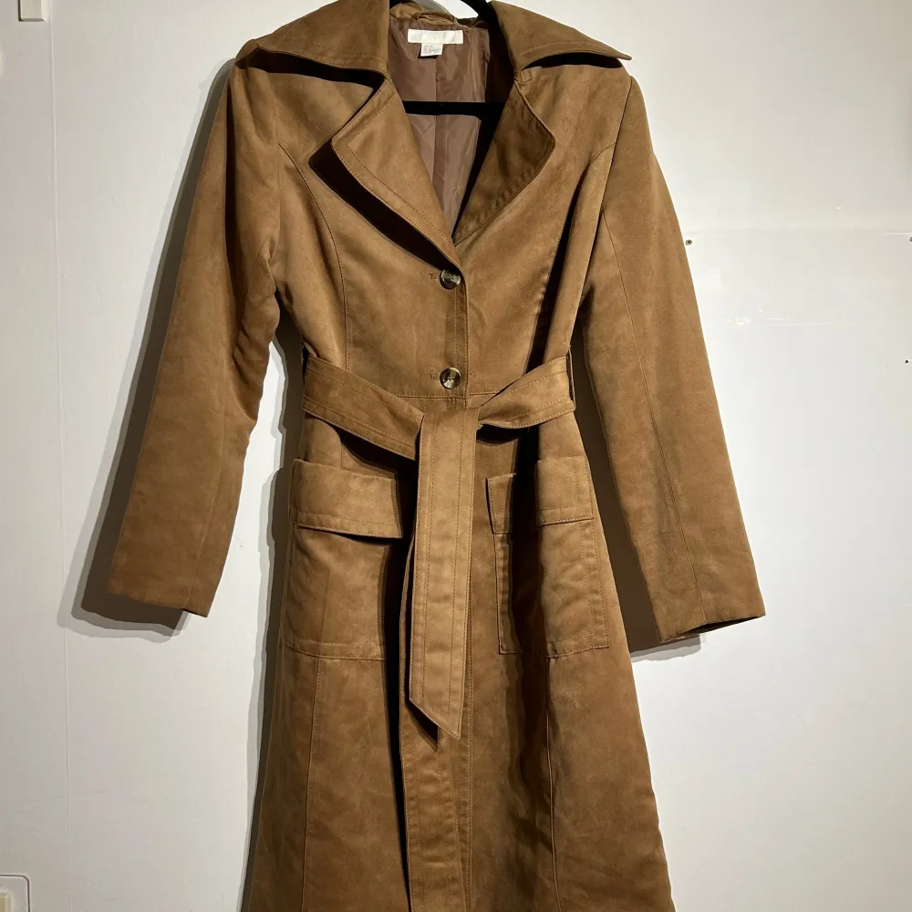 Säljer en snygg brun trenchcoat i mocka med knappar och bälte i midjan. Jackan har en klassisk krage. Den är i mycket bra skick och ger en stilren look.. Jackor.