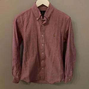 Rosa skjorta från Ralph Lauren - Säljer en snygg röd skjorta från Ralph Lauren i storlek S. Skjortan är i slim fit och har långa ärmar med knappar. Den är tillverkad i ett stretchigt oxfordmaterial och har en broderad logga på bröstet. Perfekt för både vardag och fest! Pris kan diskuteras vid snabb affär, skriv privat vid minsta fundering👍🏻