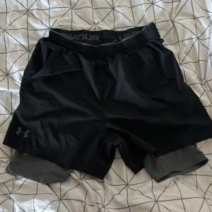 Säljer ett par svarta träningsshorts från Under Armour. De är i storlek L och har en loose passform. Shortsen är gjorda av ett lätt och bekvämt material med HeatGear-teknologi, perfekt för intensiva träningspass. De har en elastisk midja och en diskret logga på benet.