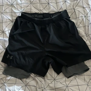 Svarta träningsshorts från Under Armour - Säljer ett par svarta träningsshorts från Under Armour. De är i storlek L och har en loose passform. Shortsen är gjorda av ett lätt och bekvämt material med HeatGear-teknologi, perfekt för intensiva träningspass. De har en elastisk midja och en diskret logga på benet.
