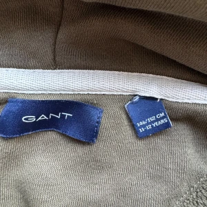 Olivgrön hoodie från GANT - Säljer en snygg olivgrön hoodie från GANT. Den har en klassisk design med en känguruficka och GANT-loggan broderad på bröstet. Perfekt för en avslappnad stil och passar bra till både vardag och skola.