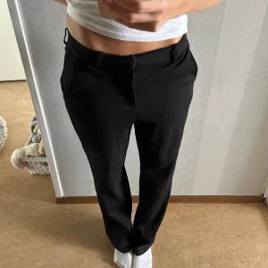 Svarta kostymbyxor - Säljer ett par stilrena svarta kostymbyxor. De är lågmidjade och har en klassisk rak passform. Perfekta för både jobb och festliga tillfällen. Byxorna har en dragkedja och knappar framtill för enkel stängning.