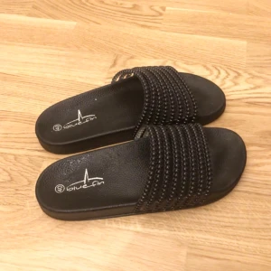 Svarta sandaler från Blue Fin - Säljer ett par svarta tofflor i storlek 40. De har en cool design med flera rader av pärlliknande detaljer på remmen.