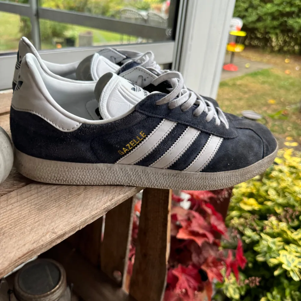 Säljer ett par klassiska Adidas Gazelle sneakers i mörkblå mocka med vita detaljer. Skorna har de ikoniska tre ränderna på sidan och texten 'GAZELLE' i guld. De är i bra skick med lite slitage på sulorna. Perfekta för både vardag och fest! Ingår även mörkblåa skosnören!. Skor.