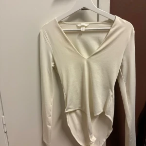 Vit långärmad body från H&M - Säljer en vit/creme långärmad body från H&M i storlek S. Toppen har en v-ringad design och är tillverkad i ett mjukt och bekvämt material. 