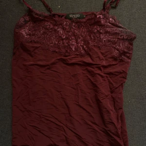 Vinröd spetslinne från Soaked in Luxury - Snyggt vinrött linne från Soaked in Luxury med spetsdetaljer upptill. Perfekt för en kväll ute eller som ett lager under en kavaj. Linnet har tunna axelband och är tillverkat i ett mjukt material som känns skönt mot huden.🍷❤️