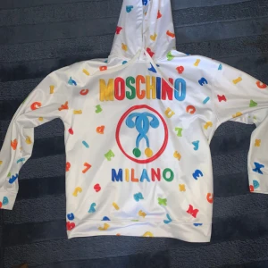 Färgglad hoodie från Moschino - Säljer en supercool vit hoodie från Moschino med färgglada bokstäver och logga. Perfekt för att sticka ut i mängden! Den har en bekväm passform och är gjord i mjukt material. Passar perfekt för både vardag och festliga tillfällen. 🧡💙💚