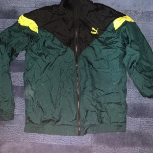 Grön och svart vindjacka från Puma - Säljer en snygg grön och svart vindjacka från Puma med gula detaljer. Jackan har dragkedja och är perfekt för höst och vår. Den är i bra skick och har en normal passform. Perfekt för träning eller vardagsbruk!