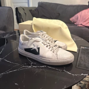 Vita sneakers från Axel Arigato - Säljer ett par stilrena vita sneakers från Axel Arigato. De har en snygg svart stjärndetalj på sidan och är i bra skick. Perfekta för både vardag och fest. Skorna har snörning och en bekväm passform.