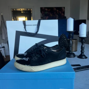 Svarta sneakers LANVIN - Säljer ett par snygga svarta Lanvin sneakers med glansiga lackdetaljer på tån och hälen. Skorna har en vit sula och klassisk snörning. Perfekta för både vardag och fest! De är i bra skick och redo för nya äventyr.