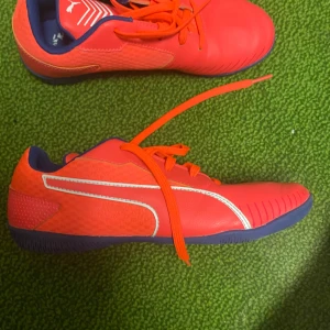 Röda fotbollsskor från Puma - Säljer ett par snygga röda fotbollsskor från Puma. De har en cool design med vita detaljer och blå insida. Skorna är i bra skick och perfekta för inomhusfotboll. Snörningen ger en bra passform och de är riktigt bekväma att spela i. Passa på att köpa dessa grymma skor!