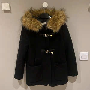 Svart kappa med fuskpäls från Eva Rosa - Säljer en elegant svart kappa från Eva Rosa i storlek M. Den har en mysig fuskpäls runt huvan och stängs med snygga spännen och dragkedja. Perfekt för kyliga höst- och vinterdagar. Kappan har långa ärmar och praktiska fickor framtill. Ett måste för garderoben! 🖤