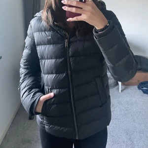 Svart dunjacka från Tommy Hilfiger - Säljer en svart dunjacka från Tommy Hilfiger i mycket bra skick. Jackan har en dragkedja framtill och är perfekt för kalla dagar. Den är långärmad och har en klassisk pufferstil med märkets logga på bröstet och ärmen. Supermysig och varm för vintern!