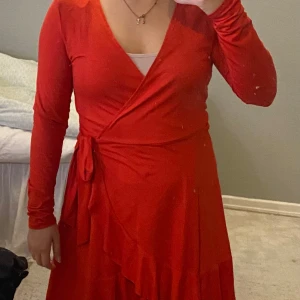 Julklänning - Snygg röd omlottklänning med långa ärmar och knytband i midjan. Perfekt för höst och vinter, med en härlig volangdetalj som ger extra stil. Klänningen har en v-ringad design och är i bra skick. Passar både till vardag och fest! ( det är spegeln som är smutsig)