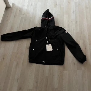 Svart vindjacka med huva - Säljer en svart vindjacka med huva och snygga ränder i vitt och rött. Jackan har en dragkedja framtill och praktiska fickor med knappar. Perfekt för blåsiga dagar och håller dig stilren. Jackan är i mycket bra skick och passar perfekt för höst och vår. Kan gå ner lite i pris!! Jackan är äkta också!!!