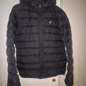 Svart pufferjacka med huva - Säljer en svart pufferjacka från Lyle & Scott i mycket bra skick. Jackan har en praktisk huva och dragkedja framtill. Perfekt för kyliga höst- och vinterdagar. Den är långärmad och har en liten gul logga på bröstet. Superbekväm och stilren!