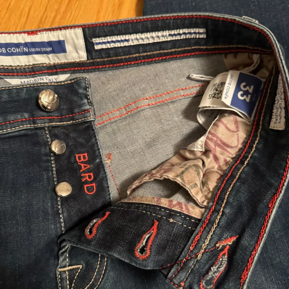 Säljer ett par snygga mörkblå jeans från Jacob Cohen i storlek 33. De har en klassisk femficksdesign med röda sömmar och en cool orange läderpatch med logga baktill. Jeansen har knappar istället för dragkedja och en broderad detalj på myntfickan. Perfekta för både vardag och fest! Bra skick förutom en defekt utpekad med röd pil.. Jeans & Byxor.