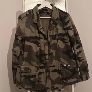 Camouflagejacka från Gina Tricot - Säljer en snygg camouflagejacka från Gina Tricot i 100% bomull. Jackan har en cool militärstil med flera fickor och dragkedja. Perfekt för höst och vår, och den har långa ärmar. Passar bra till både vardag och äventyr!  Köpt ett tag sedan men har knappast använts. 