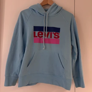 Ljusblå hoodie från Levi's - Säljer en snygg ljusblå hoodie från Levi's i bra skick. Den har en stor ficka fram och en klassisk logga i blått, rött och rosa. Perfekt för en avslappnad stil och passar till alla årstider. Skön och mjuk, perfekt för vardagsbruk!