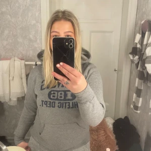 Grå hoodie med blå text - Mysig grå hoodie med blå text 'Athletic Dept' på bröstet. Perfekt för en avslappnad stil och håller dig varm under kyliga dagar. Har en klassisk känguruficka framtill och en bekväm huva. Passar perfekt för vardagsbruk eller en chill dag hemma.