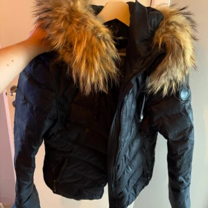 Svart dunjacka från Hollies - Säljer en snygg svart dunjacka från Hollies i storlek 38. Jackan har en fluffig pälsdetalj på huvan och är perfekt för kalla vinterdagar. Den är i mycket bra skick och har en dragkedja framtill. Supermysig och stilren för vintern! ❄️