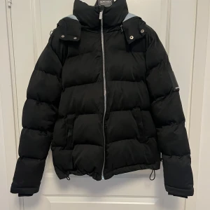 Svart pufferjacka från Manière De Voir - Säljer en snygg svart pufferjacka från Manière De Voir i storlek M. Jackan har en dragkedja framtill och en huva med grå detaljer. Perfekt för kalla dagar med sin tjocka vaddering. Den är i bra skick med en liten skada på insidan men redo för vintern! ❄️