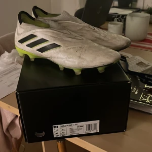 Adidas Copa Pure+ FG Fotbollsskor - Säljer ett par Adidas Copa Pure+ FG fotbollsskor i vitt med svarta och gröna detaljer. Skorna är i bra skick och perfekta för fotboll på gräsplaner. De har en stilren design utan snörning för en tight passform. Perfekta för dig som vill ha både stil och funktion på planen!