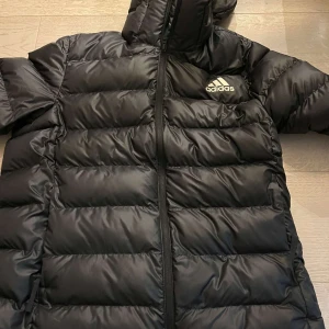 Svart pufferjacka från Adidas - Säljer en svart pufferjacka från Adidas i storlek L. Jackan är perfekt för kalla dagar med sitt quiltade mönster och långa ärmar. Den har en praktisk dragkedja framtill och en huva för extra värme. Perfekt för vintern eller hösten!