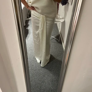 Maxikjol från Mango - Säljer en maxikjol i glansigt material och mönster.  Vit/krämvit med en detalj framtill samt slits. Dragkedja på sidan 