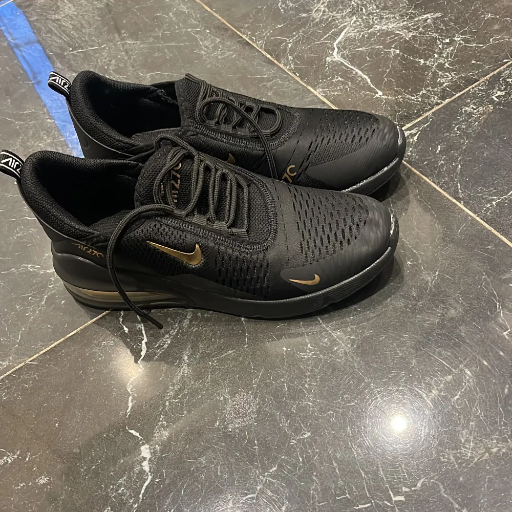 Säljer ett par svarta Nike Air Max 270 i mycket bra skick. De har en snygg design med guldiga detaljer och en bekväm passform. Perfekta för både vardag och träning. Storleken är 42 i herrstorlek.. Skor.