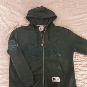 Mörkgrön hoodie från Pull&Bear - Säljer en mörkgrön hoodie från Pull&Bear i samarbete med Starter. Den har en dragkedja framtill och en stor logga på bröstet. Skick 8/10. 