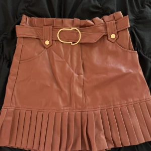 Brun kjol från Zara - Snygg brun kjol från Zara i skinnimitation med plisserad nederdel och ett brett bälte med guldfärgat spänne. Perfekt för höst och vår, och kan stylas både upp och ner beroende på tillfälle. Kjolen har en modern och chic look som passar till många olika outfits.