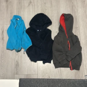 Tre mysiga barnjackor  - Säljer tre barnjackor i olika stilar. En blå fleecejacka med dragkedja, perfekt för vår och höst. En svart teddyjacka med huva, supermjuk och varm. En grå hoodie med röd insida och dragkedja, cool och bekväm för vardagsbruk.