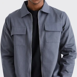 Grå overshirt från MAN - Säljer en stilren grå overshirt från MAN i mycket bra skick. Den har en dragkedja framtill och två praktiska bröstfickor med lock. Perfekt för höst och vår, och funkar både till vardags och fest. Passformen är normal och materialet känns slitstarkt och bekvämt.