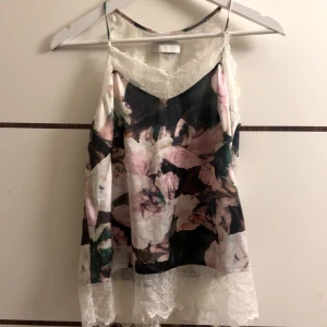 Blommig linne med spetsdetaljer - Säljer ett superfint linne med blommigt mönster i rosa och gröna toner. Det har tunna axelband och vackra spetsdetaljer längs kanterna. Perfekt för sommardagar eller en festlig kväll. Passar bra till jeans eller kjol!