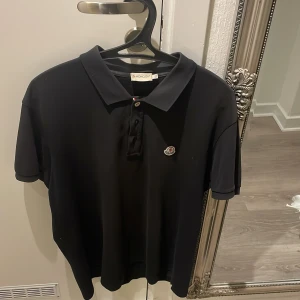 Svart pikétröja från Moncler ÄKTA - Säljer en stilren svart pikétröja från Moncler i mycket bra skick. Tröjan har en klassisk krage och knappar framtill. Perfekt för både vardag och festliga tillfällen. Moncler-loggan är snyggt placerad på bröstet. Passar perfekt för dig som gillar en tidlös stil. Funkar för alla storlekar bara ta in i byxan.