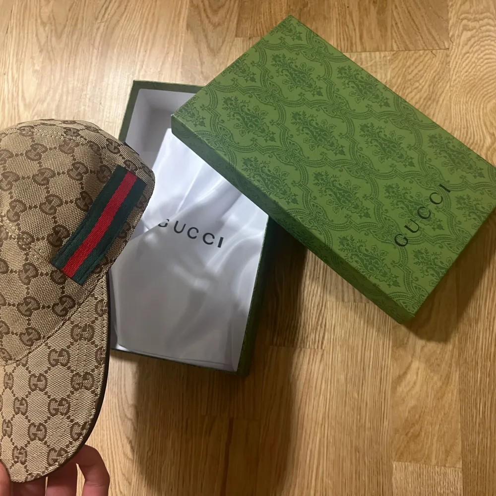 Säljer en snygg beige keps från Gucci med det klassiska GG-mönstret. Kepsen har en grön och röd randdetalj på sidan och kommer i sin originalförpackning. Perfekt för att ge din outfit en lyxig touch!. Accessoarer.