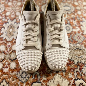 Christian Louboutin Skor - Säljer ett par vita sneakers från Christian Louboutin i skinn med coola nitar på tån. De har den klassiska röda sulan och snörning framtill. Perfekta för att ge din outfit en edgy touch! Använda men i bra skick.