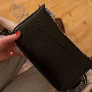 Svart kuvertväska med krokodilmönster - Snygg svart kuvertväska i skinnimitation med krokodilmönster. Perfekt för en utekväll eller fest! Väskan har en dragkedja upptill för säker förvaring av dina saker. Enkel och stilren design som passar till det mesta.