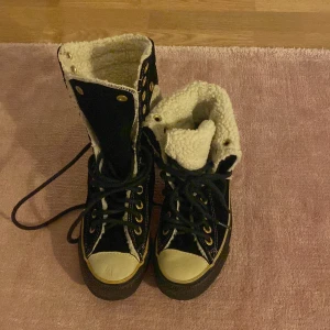 Svarta vinterkängor med foder - Säljer ett par svarta Converse-kängor i bra skick. De har en mysig, vit foder inuti som håller fötterna varma under vintern. Skorna har snörning och en klassisk Converse-logga på sidan. Perfekta för kalla dagar!